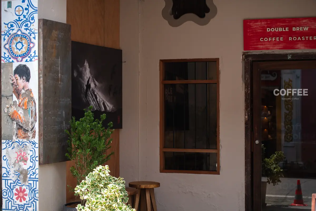 Shophouse front with a red sign above wooden doors, a plant and a stool.
