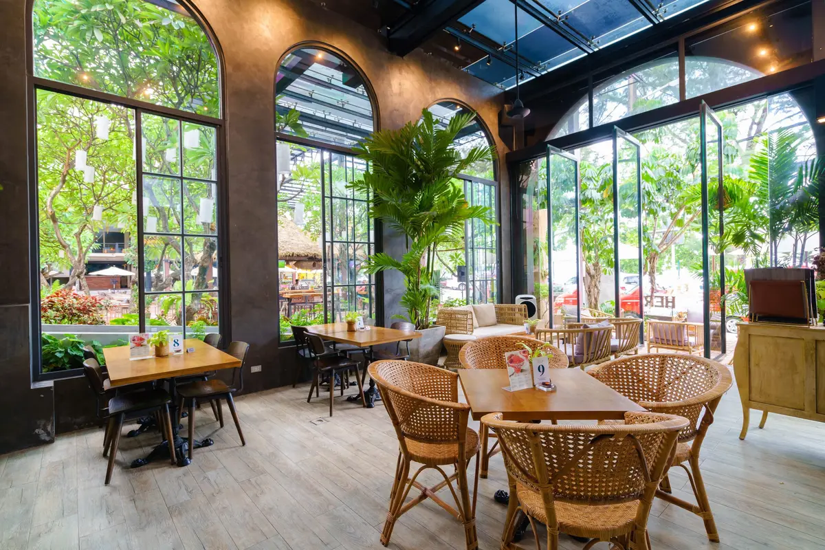 Wooden tables and rattan chairs in a bright room with tall arched windows and plants.