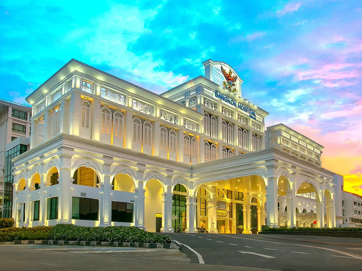 Cream-coloured hospital entrance building with arches and a crest on top, lit at dusk under a pink and blue sky.