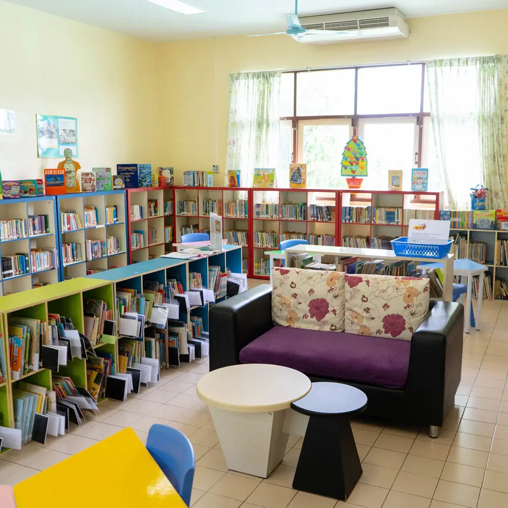 Bright school library with low shelves of picture books, a sofa and a round table by a window.