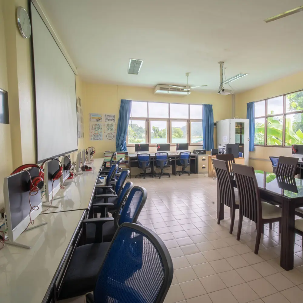 Empty classroom with wooden desks, computers along one wall and large windows.