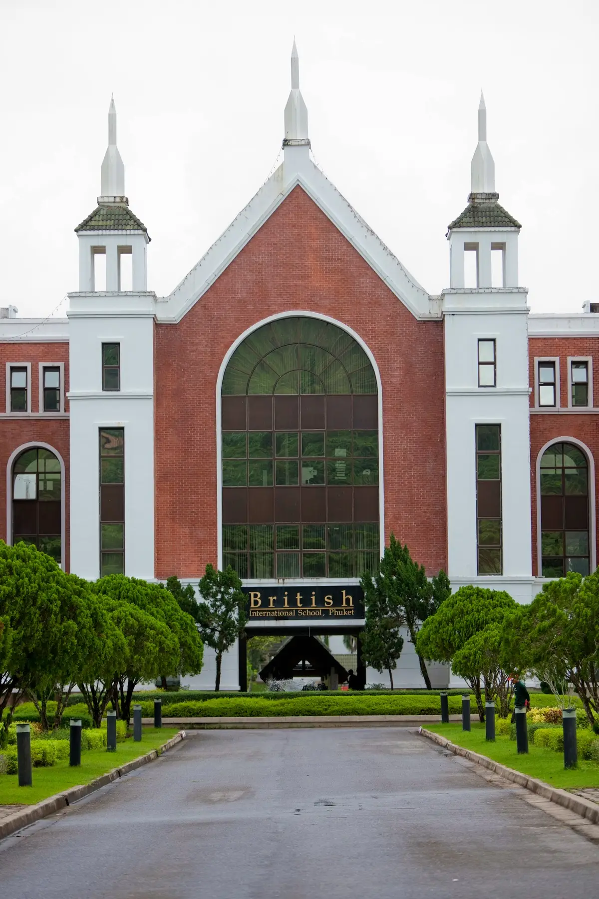 Red-brick school facade with a tall arched window and the school name on a sign among small trees.