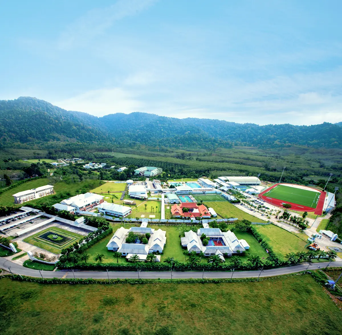 Aerial view of a spread-out school campus with white buildings, sports fields and a running track below forested hills.