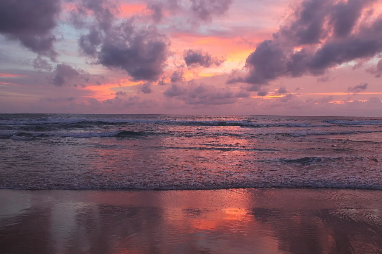 Pink and purple clouds reflected on wet sand and waves at Karon Beach at dusk.