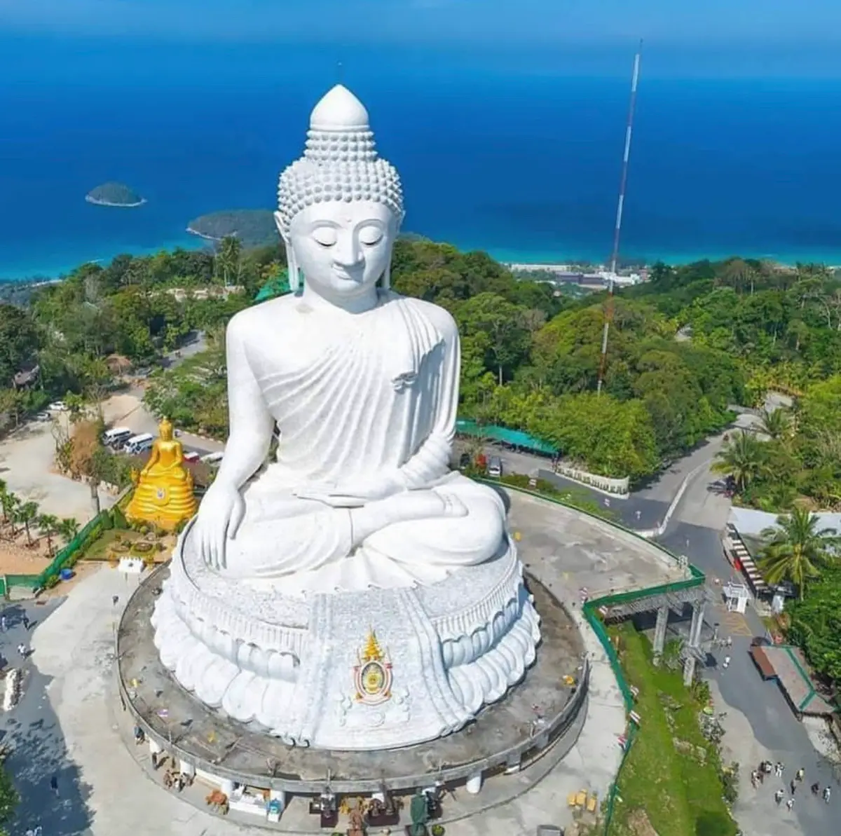 Aerial view of a large white seated Buddha statue on a hilltop with green slopes and the sea behind.