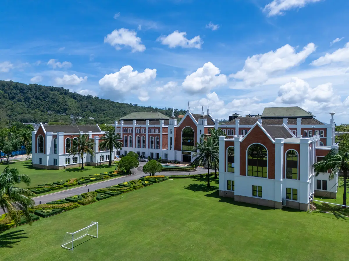 Red-brick and white school buildings with arched windows across a wide lawn with a football goal under a blue sky.