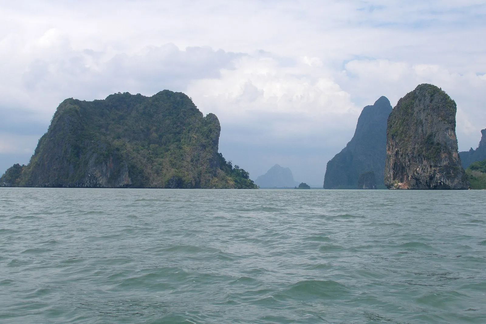Steep limestone islands rising from flat grey-green water under a cloudy sky in Phang Nga Bay.