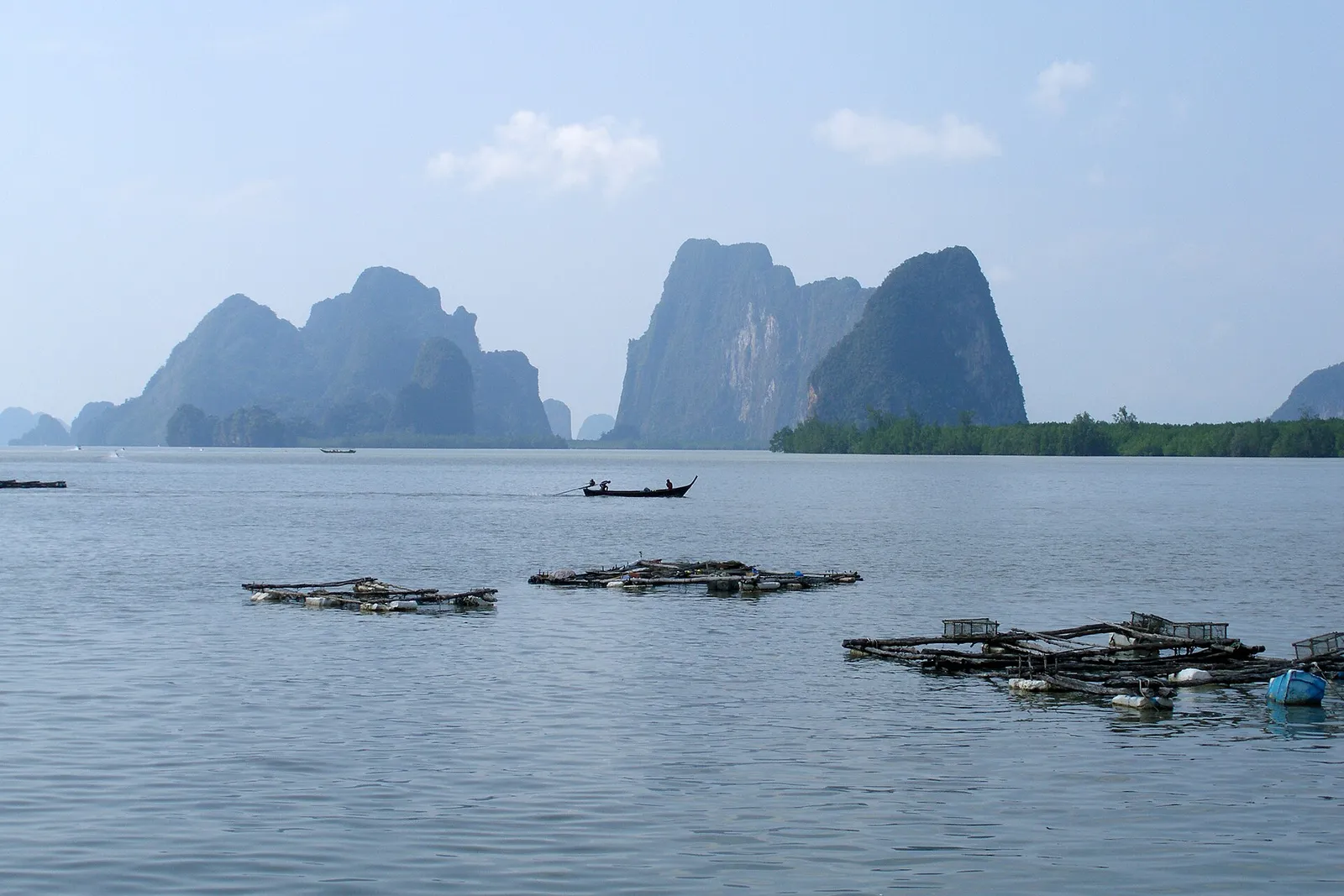 A line of limestone islands under a hazy sky across still water, with floating fish cages in the foreground.