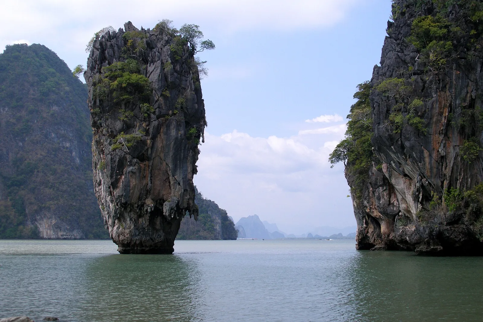 A slim limestone needle rock standing in calm green water between steep forested cliffs in Phang Nga Bay.