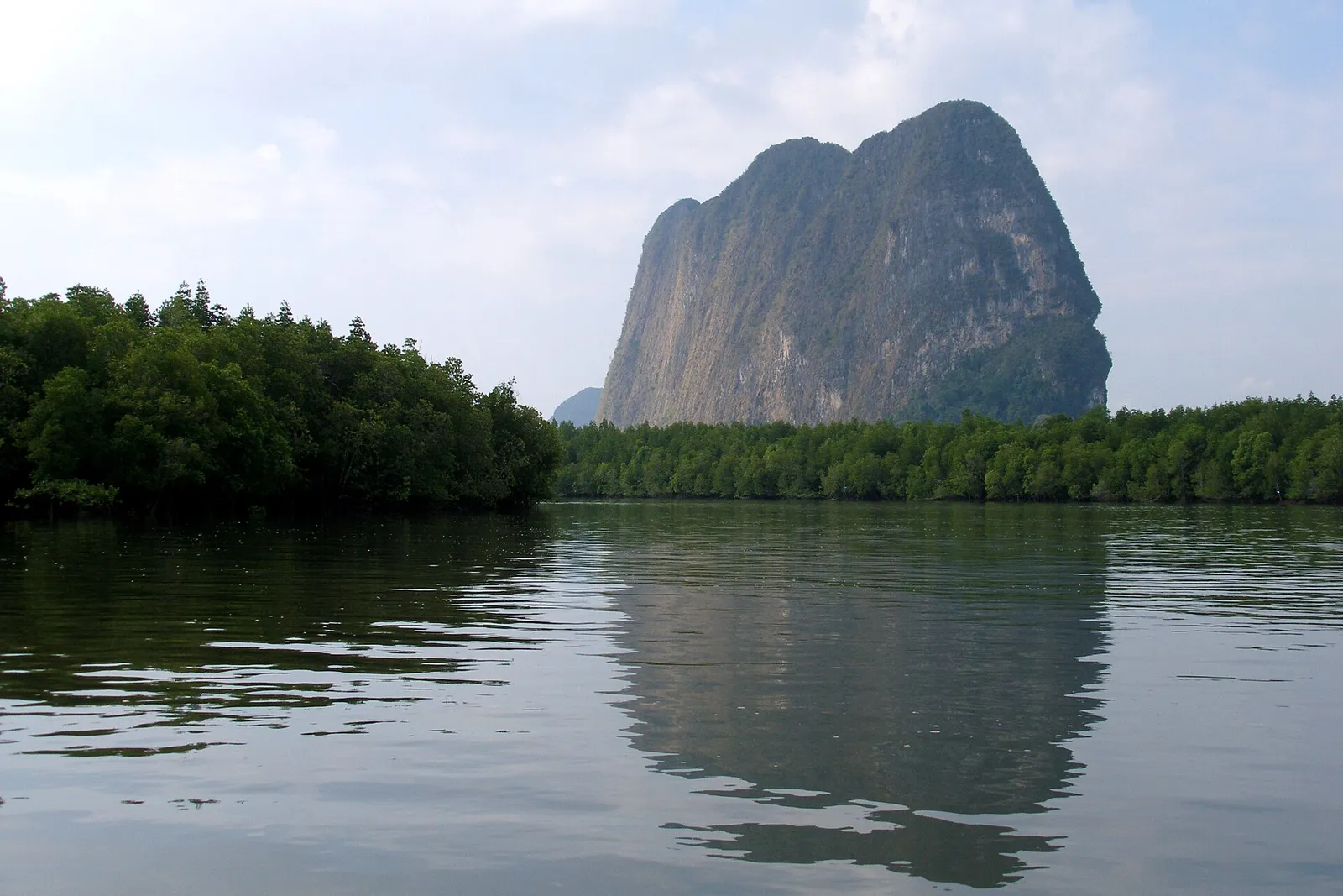 A tall limestone hill behind a green mangrove shoreline, reflected in a quiet channel.
