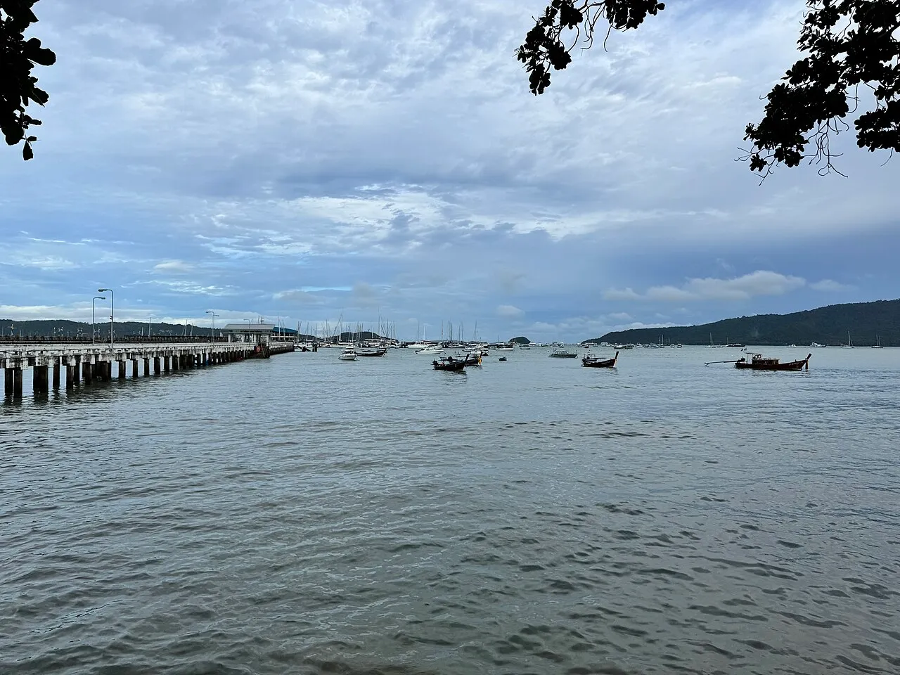 Long concrete pier on the left and moored boats scattered across a wide grey bay under a cloudy sky.