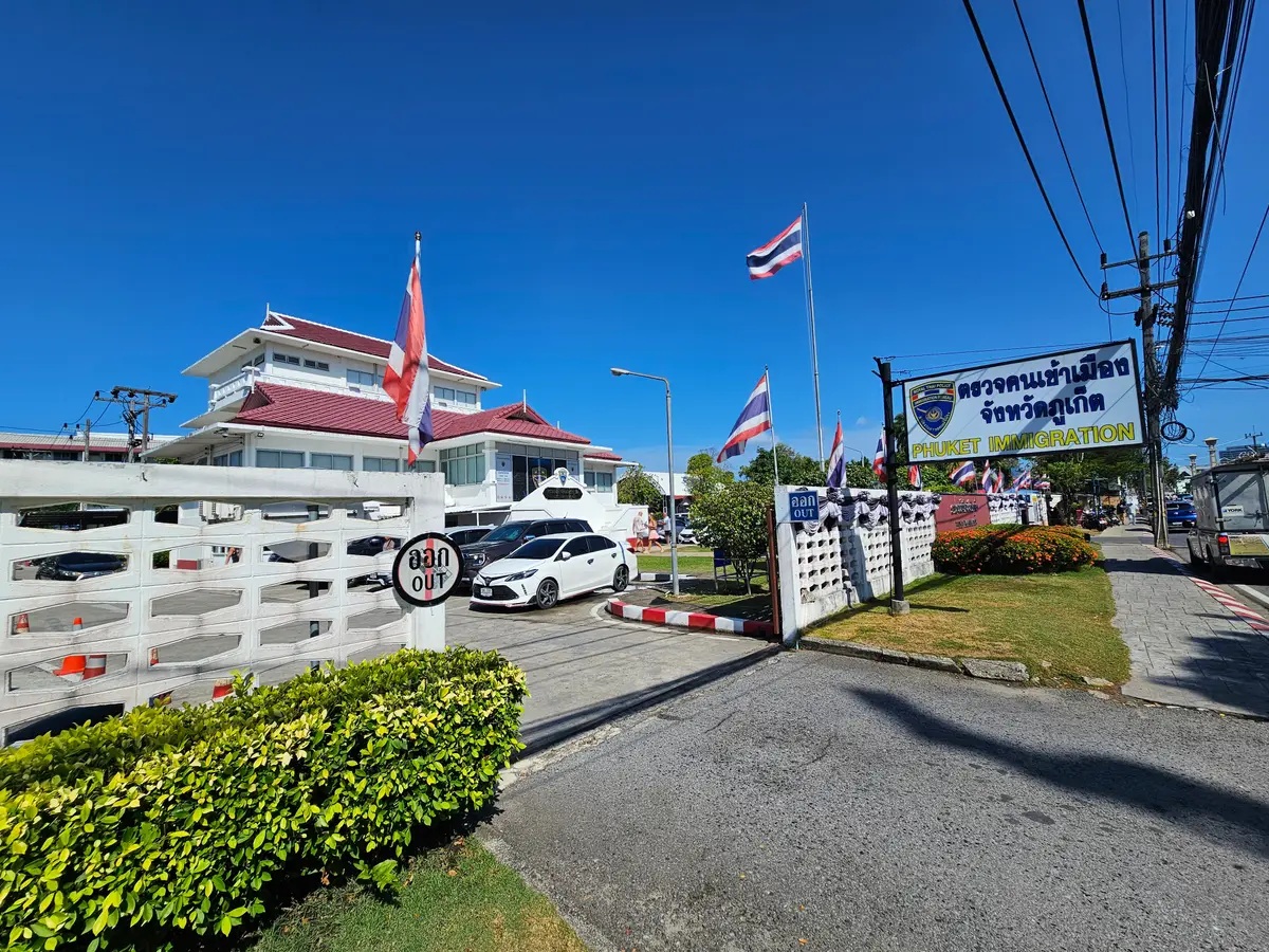 Front of the Phuket Immigration office, a white building with a red roof, flags at the entrance and a blue name sign on the white fence.
