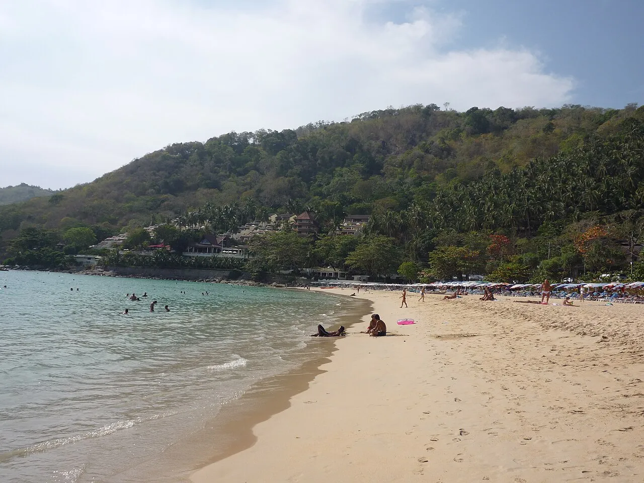 Curved sandy bay backed by forested hills with calm shallow water at Nai Harn.