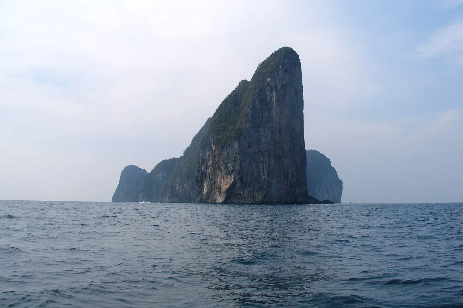 A tall limestone sea stack rising from calm open water off Ko Phi Phi Leh under a hazy sky.