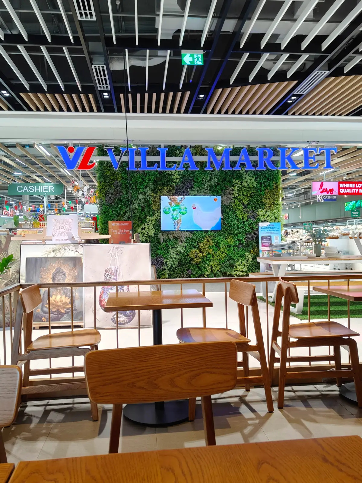 Green wall with a Villa Market sign above a cafe seating area with wooden chairs.