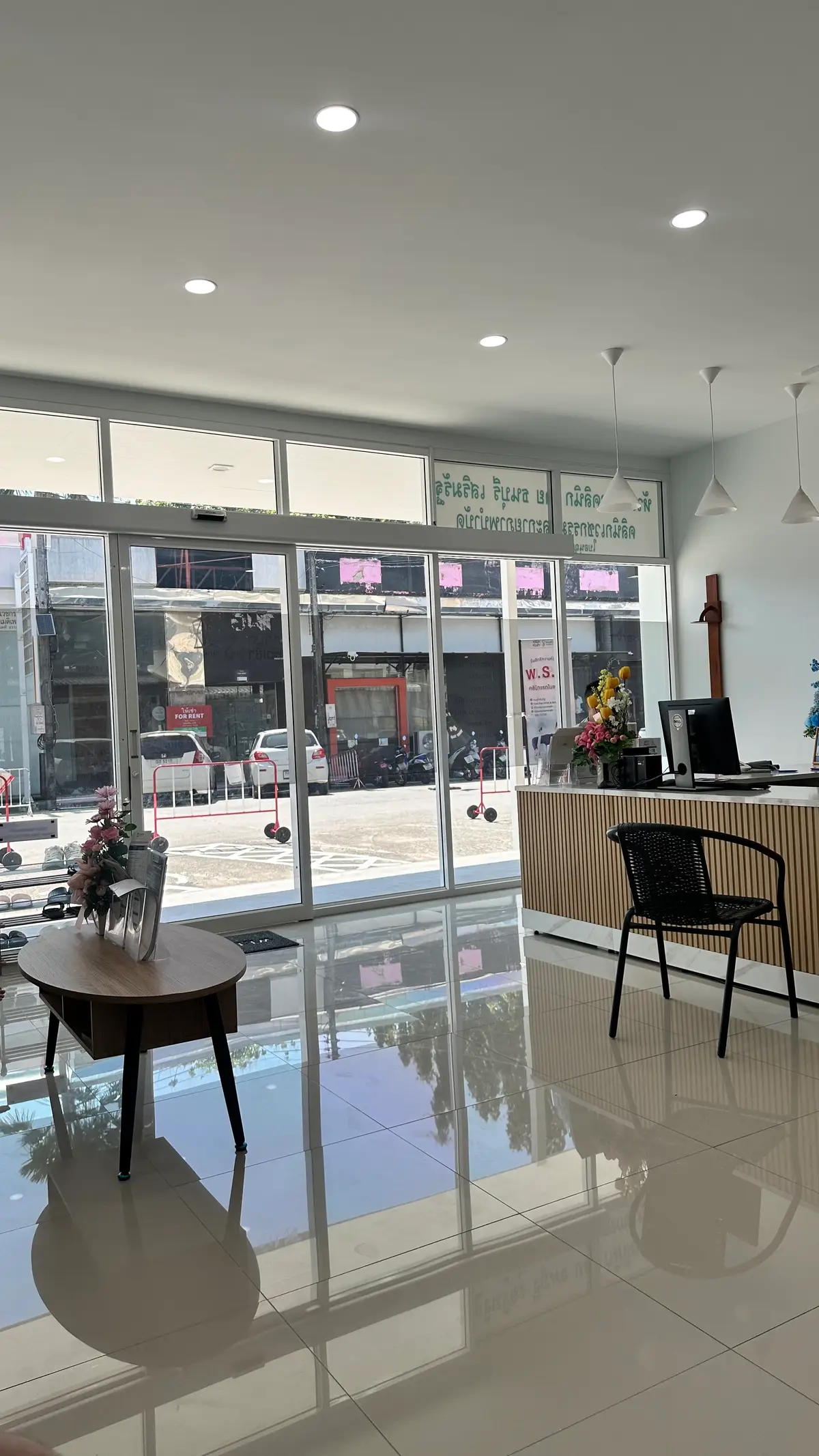 Glass entrance seen from a wooden reception counter with a black chair and a vase of pink flowers.