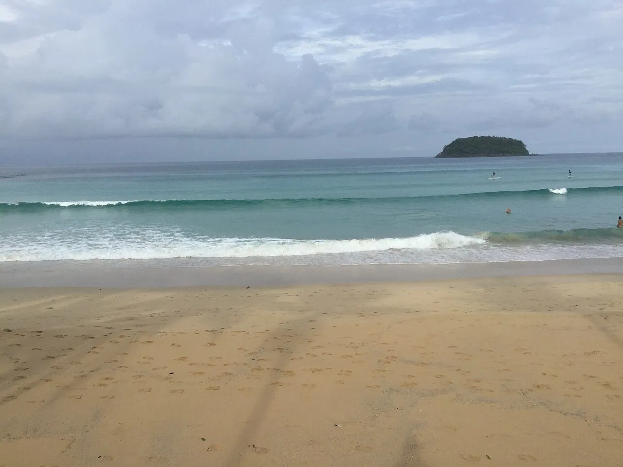 Waves rolling onto a wide sandy beach with a small green island on the horizon at Kata.