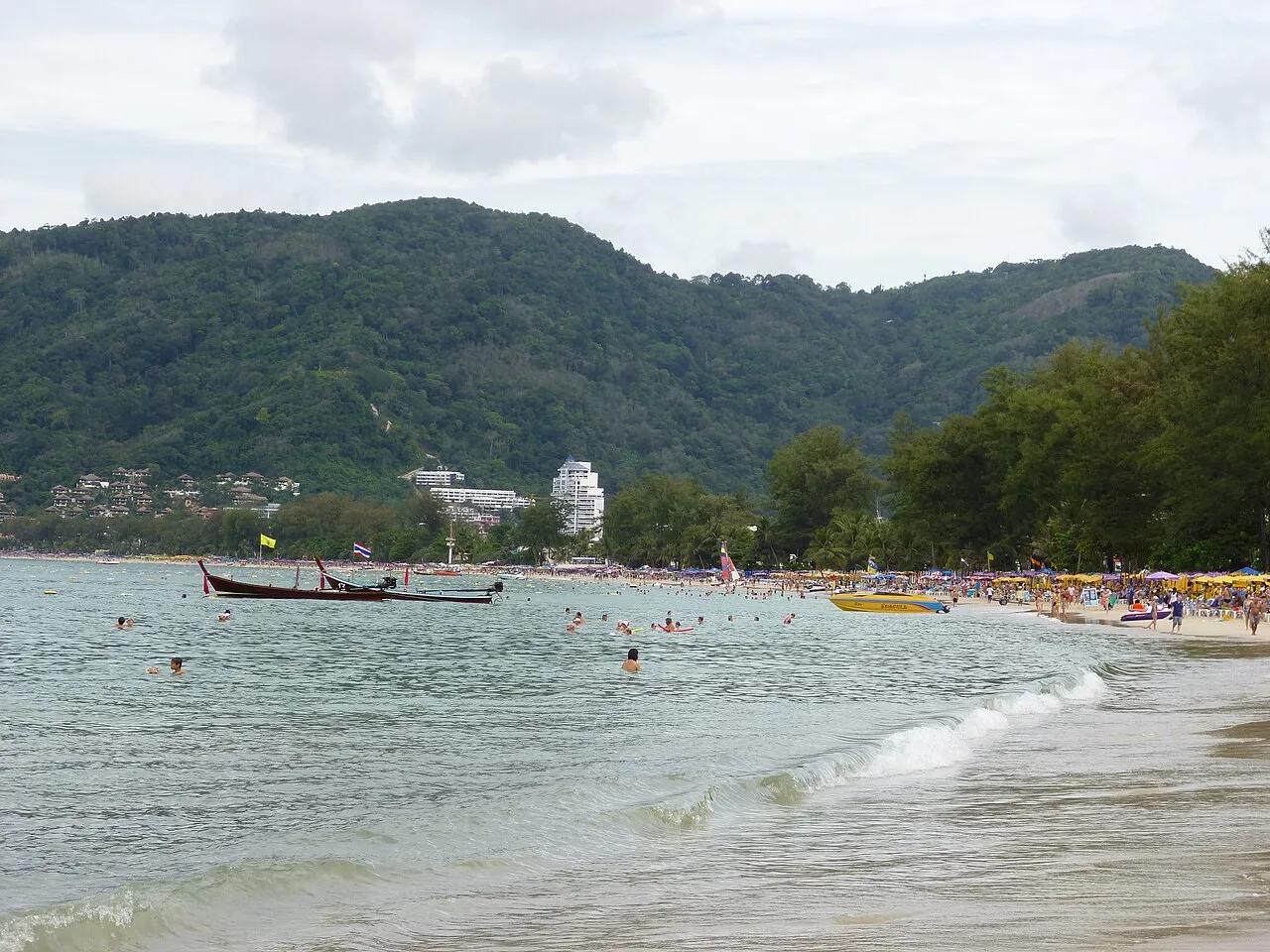 Long-tail boats on grey water in front of cloud-covered hills at Patong Bay.