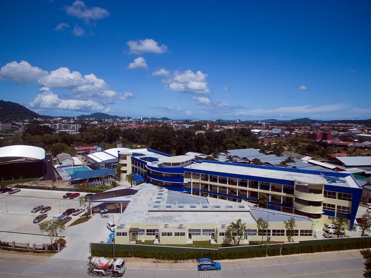 Blue and white school buildings with a car park in front, seen from a raised point with hills behind.