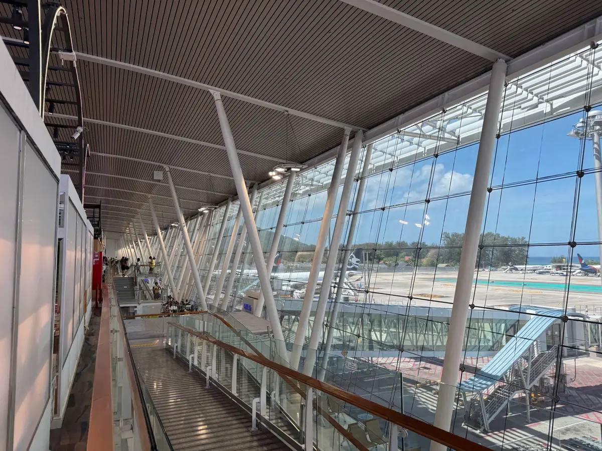Glass-walled walkway inside Phuket International Airport with escalators and a view of the aircraft apron outside.