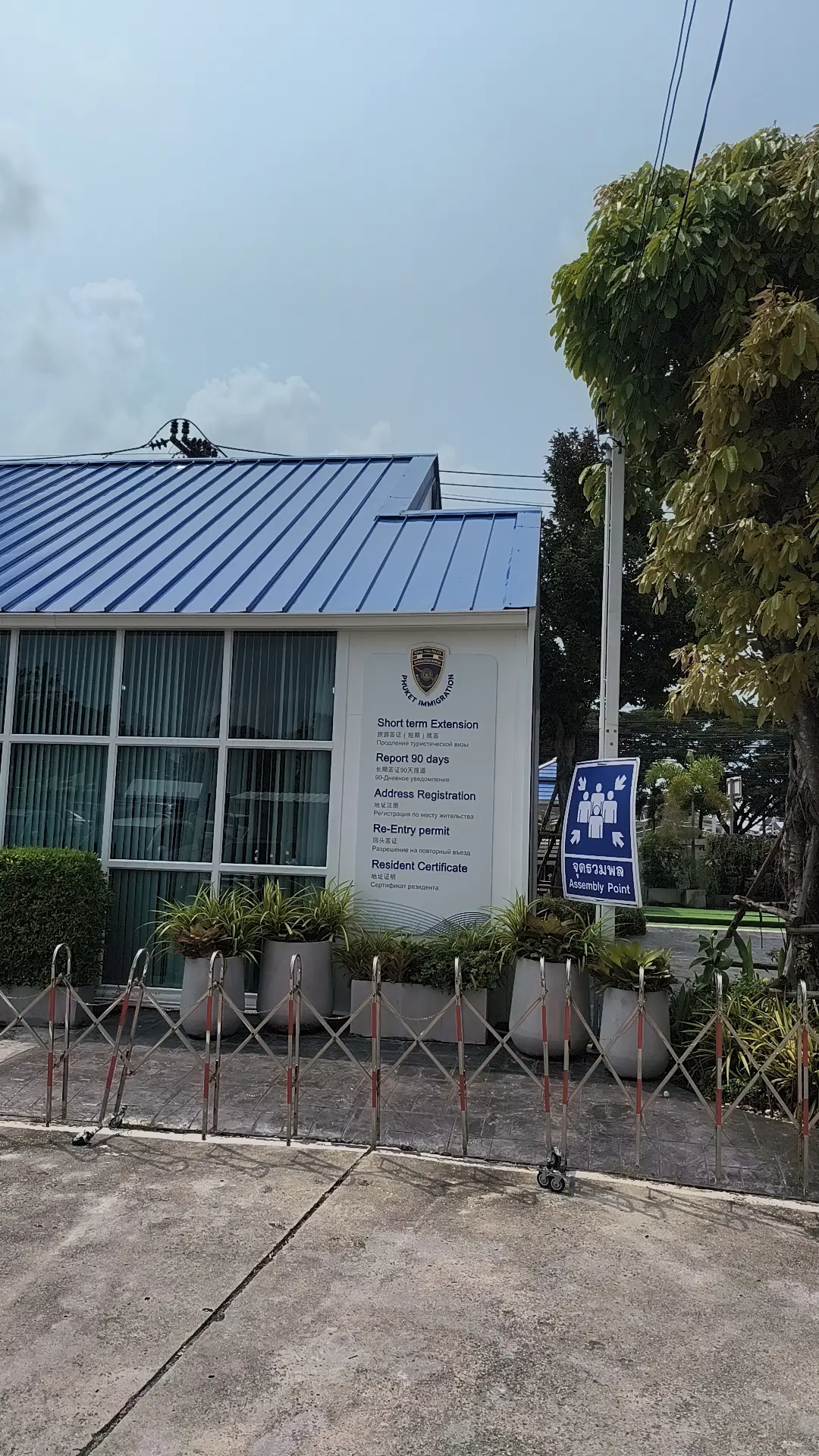 Front of a blue-roofed Phuket Immigration building with a noticeboard listing its services and a queue sign beside the entrance.