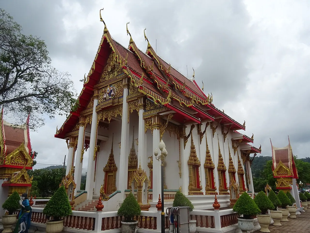 White prayer hall with a red and gold tiered roof and gilded gables under a cloudy sky at Wat Chalong.