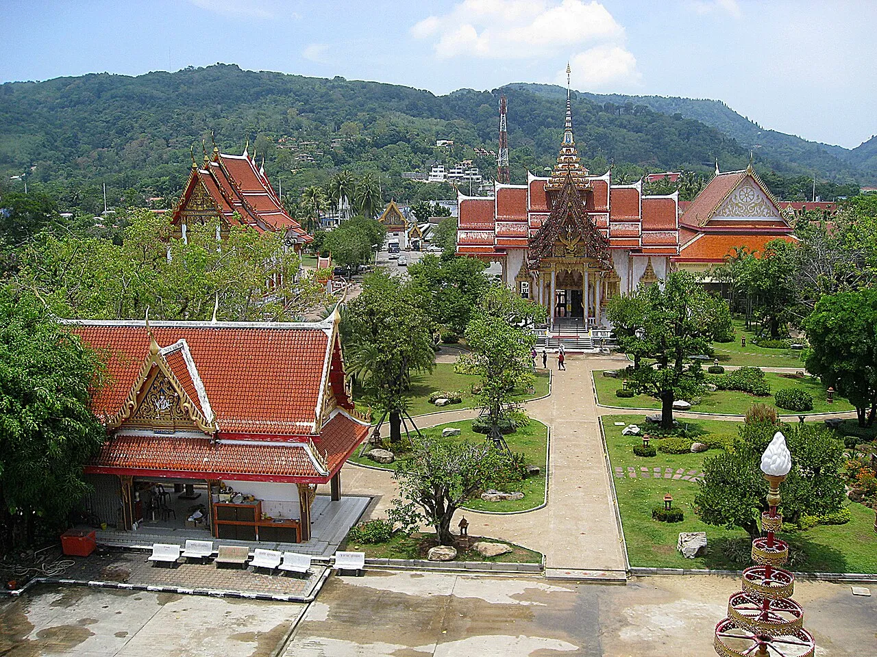 Overhead view of the Wat Chalong grounds: red-roofed halls, lawns, paths and forested hills behind.