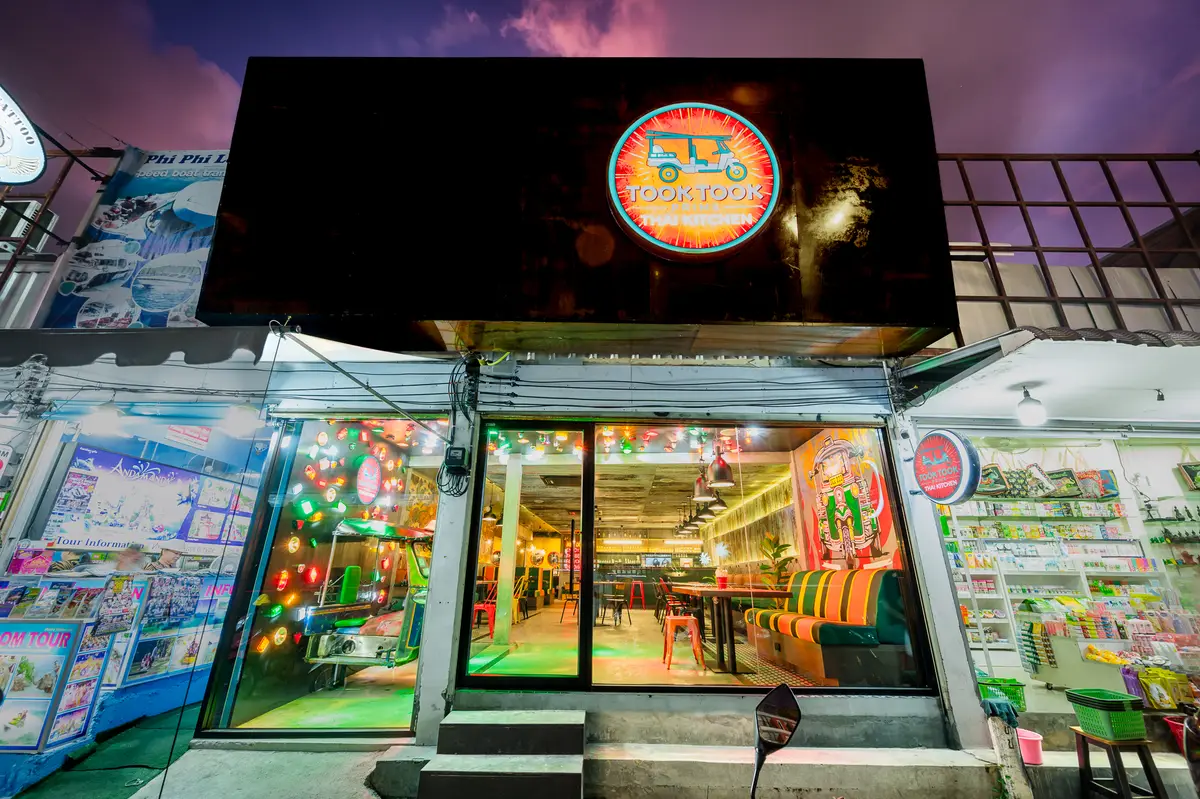 Restaurant shopfront at night with a round tuk-tuk logo sign and glass doors showing colourful tables.