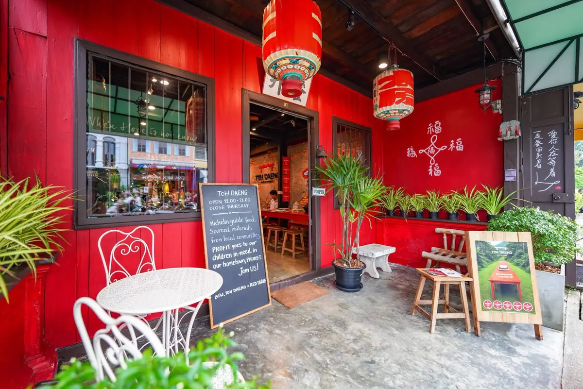 Red-painted shophouse front with a chalkboard sign, white chairs and potted plants.