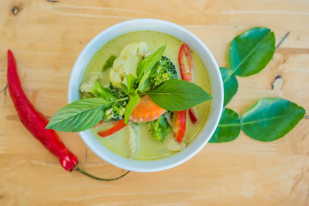 White bowl of green curry with vegetables and basil leaves on a wooden board.