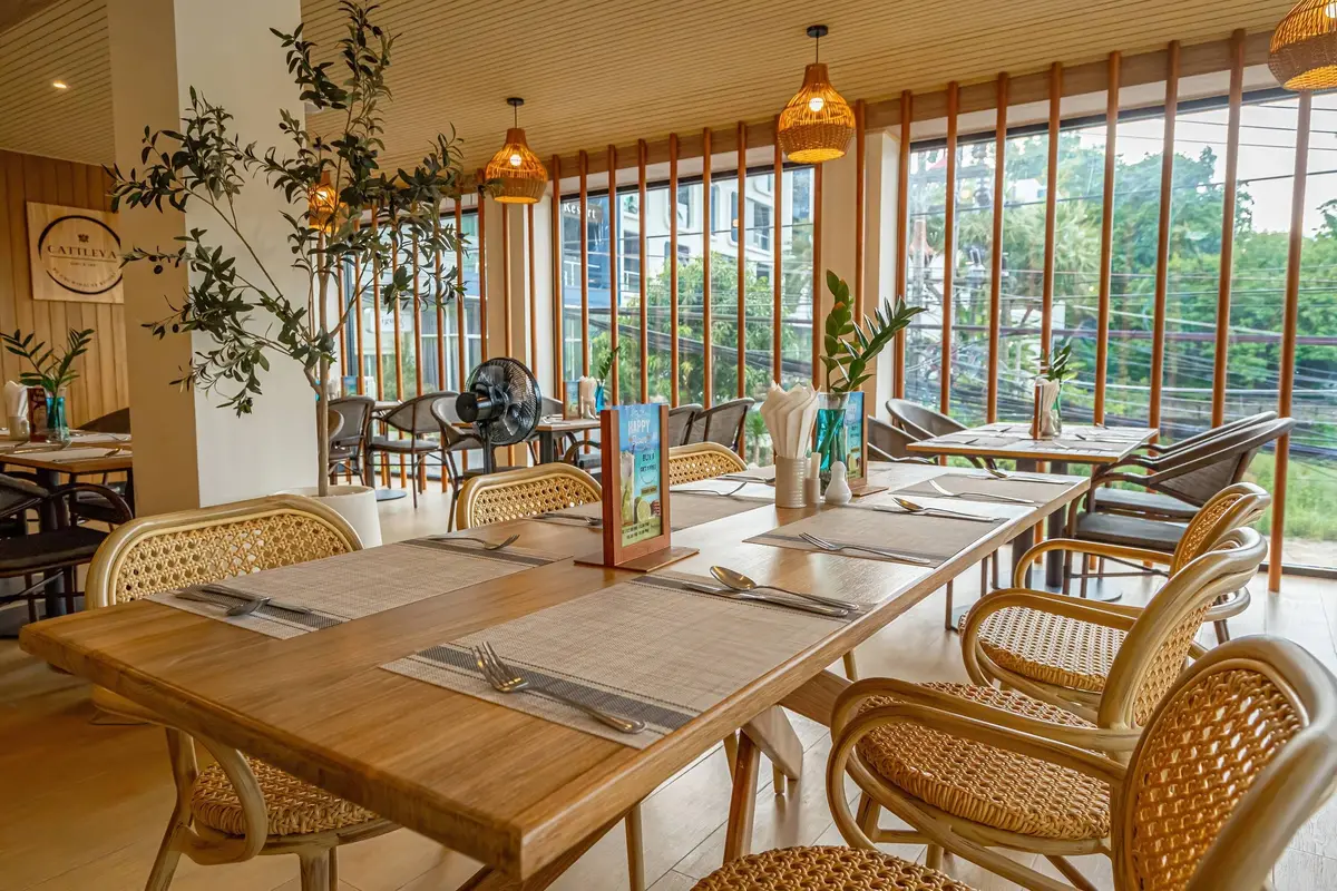 Bright dining room with wooden tables, rattan chairs and tall windows.